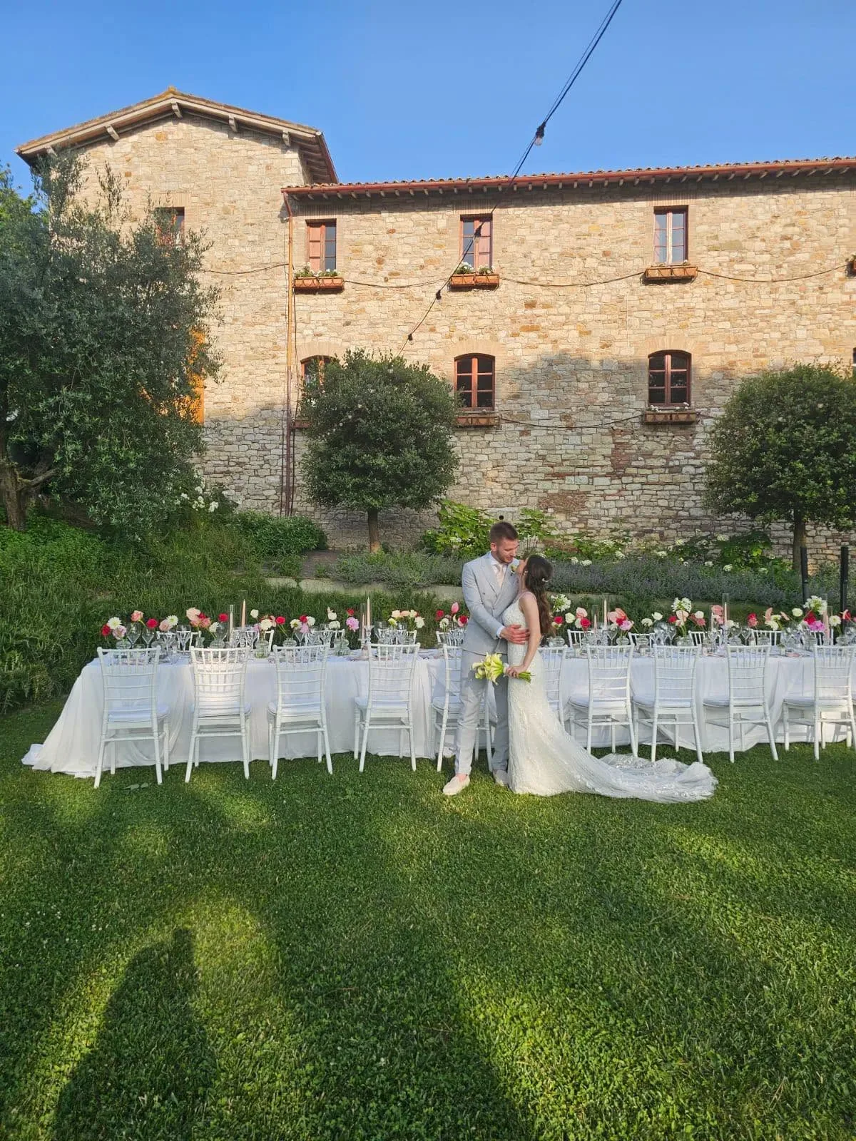 Sposi abbracciati sul prato davanti a un casale in pietra, con il tavolo imperiale allestito alle loro spalle e fiori rosa e rossi lungo tutta la tavola