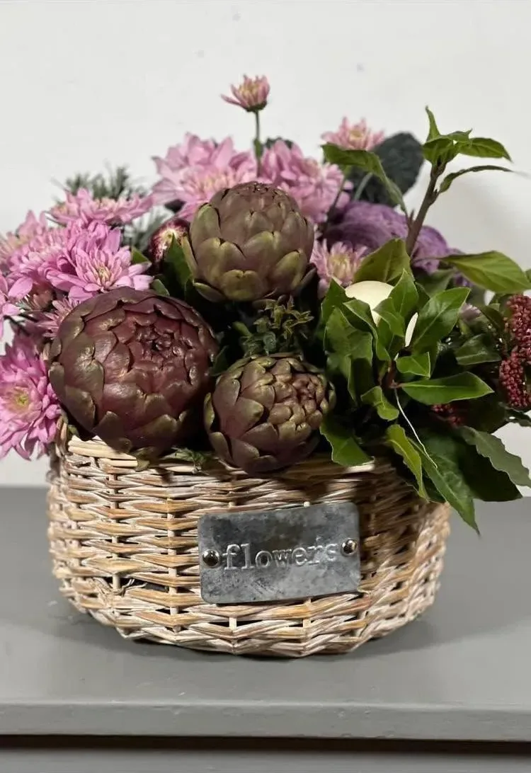 Cesto in vimini con targhetta in metallo, riempito con crisantemi rosa, carciofi e foglie di alloro