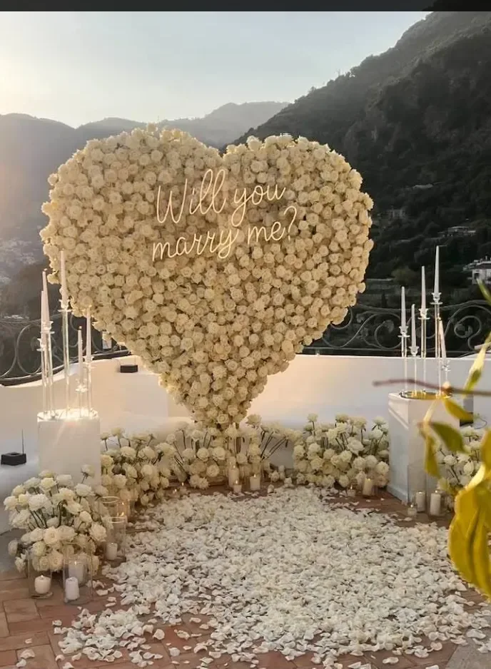 Cuore gigante di rose bianche con scritta al neon «Will you marry me?» allestito su una terrazza affacciata sulla costa, tra candele e petali a terra