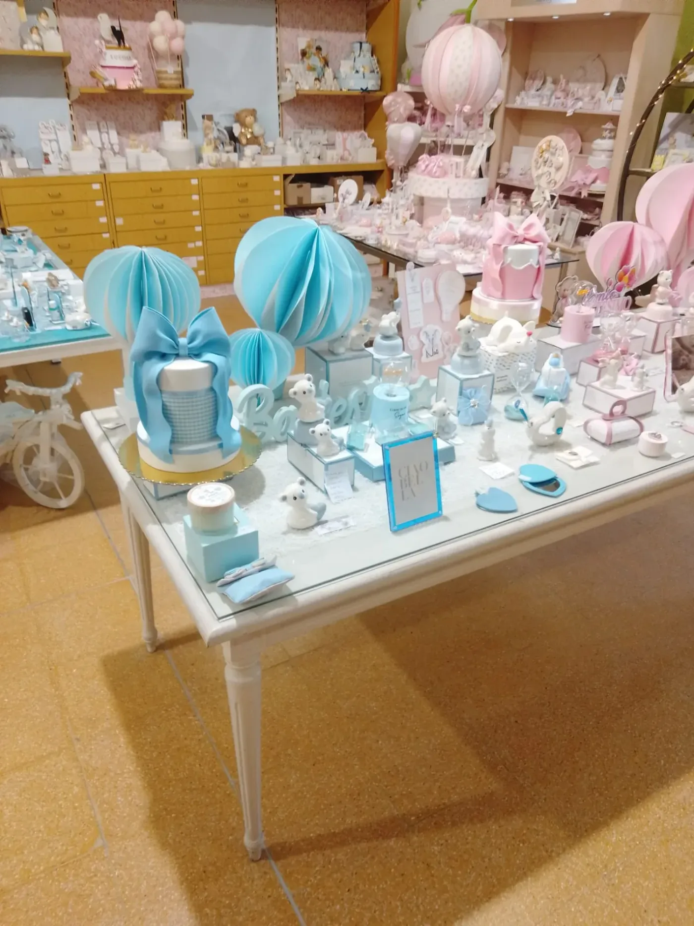 Interno de La Fioreria M. Rita Barbanera: il tavolo delle bomboniere da battesimo con torta scenografica azzurra, mongolfiere di carta rosa e celesti e scaffali di confetti e statuine