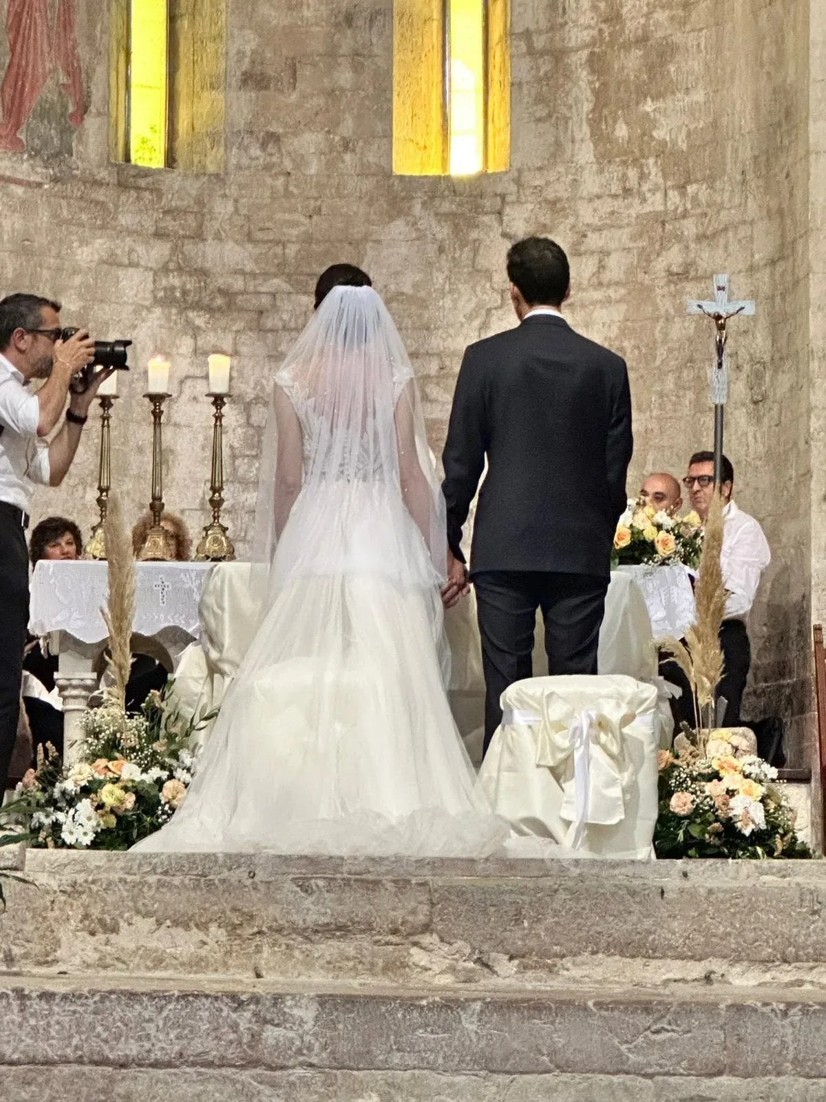 Sposi in piedi all'altare di una chiesa in pietra, tra composizioni di rose color pesca e bianche, spighe di pampas e candele sull'altare