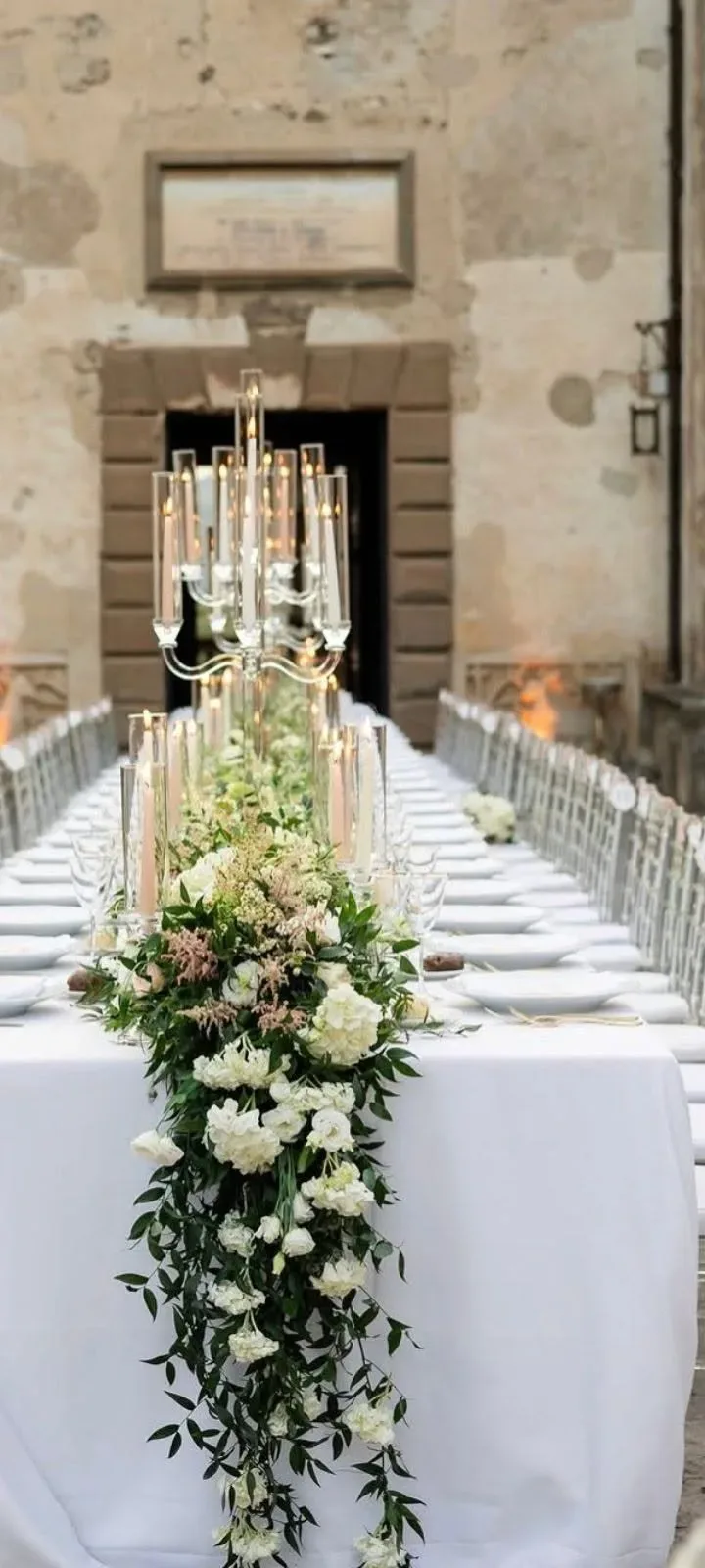 Tavolo imperiale con tovaglia bianca, candelabri di cristallo e una cascata di fiori bianchi e verde che scende dal centro tavola fino a terra, davanti al portale in pietra di un borgo